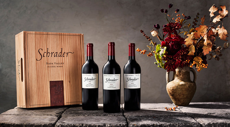 3 Bottles of Schrader Cellars Cabernet Sauvignon on cobblestones with a vase of flowers and a Schrader branded wooden box.