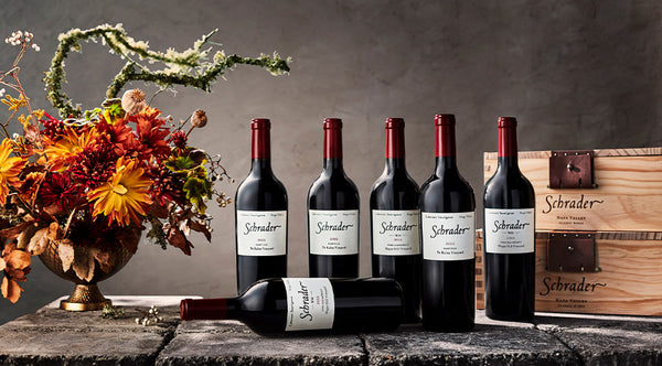 6 Bottles of Schrader Cellars Cabernet Sauvignon on cobblestones with a vase of flowers and a Schrader branded wooden box.