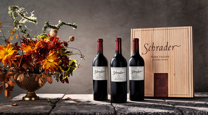 6 Bottles of Schrader Cellars Cabernet Sauvignon on cobblestones with a vase of flowers and a Schrader branded wooden box.